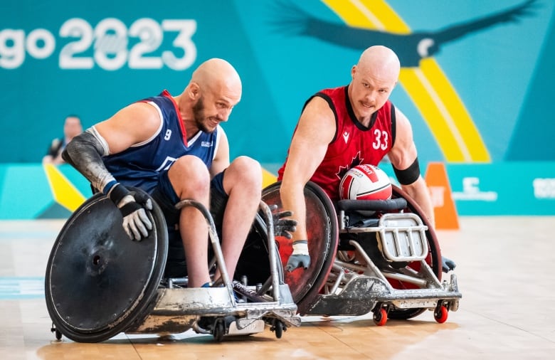 Two rugby wheelchair players- one in a blue uniform and one in red with the ball - roll beside each other during a match.