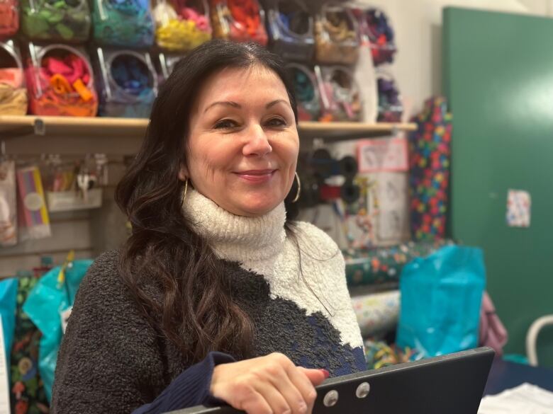 A woman stands in a toy store with a wall of gift wrapping accessories behind her.