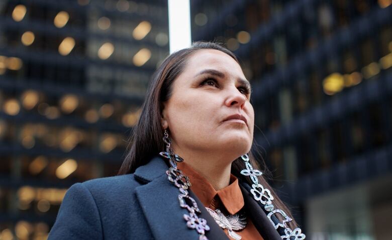 A woman with dark hair, a dark jacket and orange blouse stands in the gap of light between 2 office towers.   