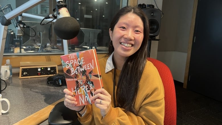 An East Asian woman holds up a book with a colourful cover while seated in a radio studio.