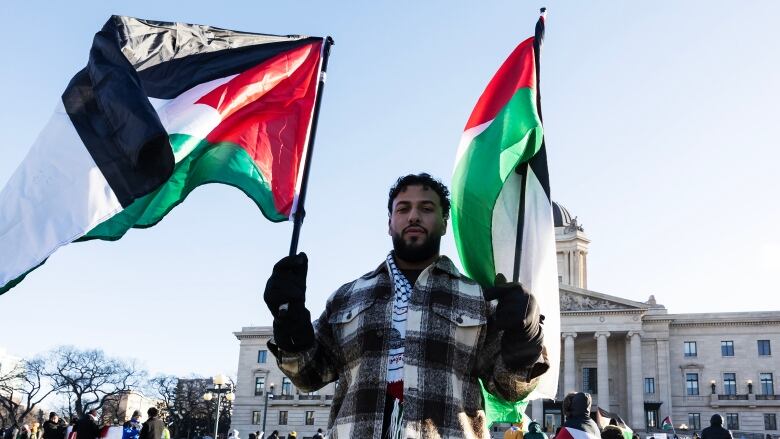 A man stands, carrying a Palestinian flag in both hands. 