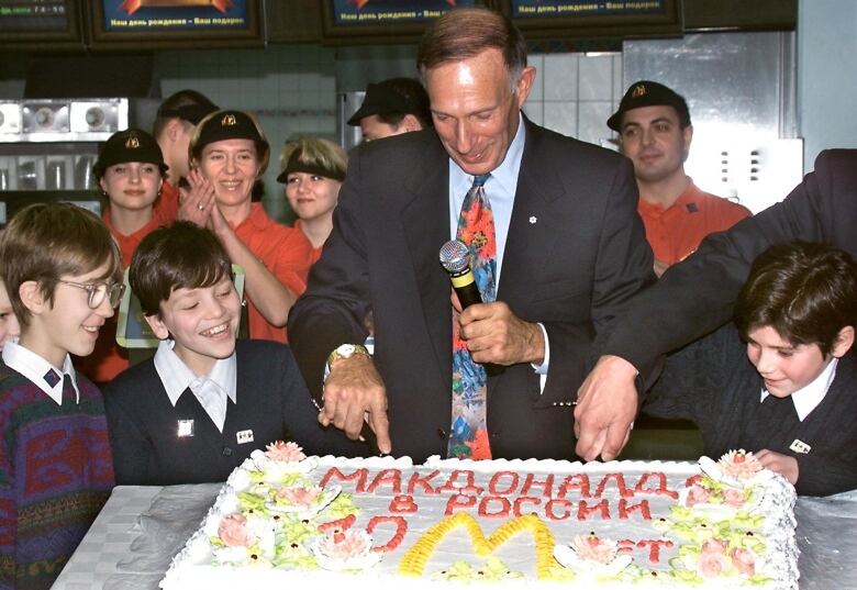 An adult cuts a cake while surrounded by children as fast food workers look on.