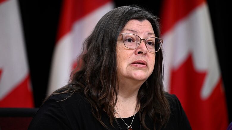 A woman with dark hair and glasses, with Canadian flags displayed in the background.