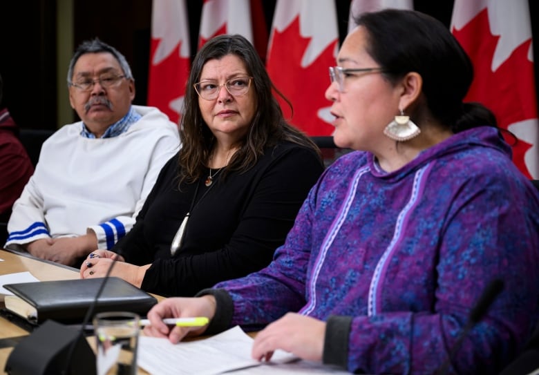 A woman sits at a table speaking into a microphone as 2 people seated beside her look on. 