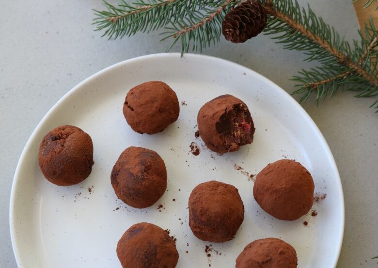 Small, rounded truffles sit on a plate. One has bite missing from it. Beside the plate is decorative branch from a pine tree.