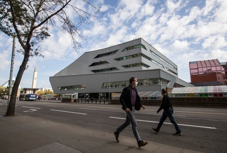 Two people walk past each other on the street in front of a square, grey building with geometic windows.