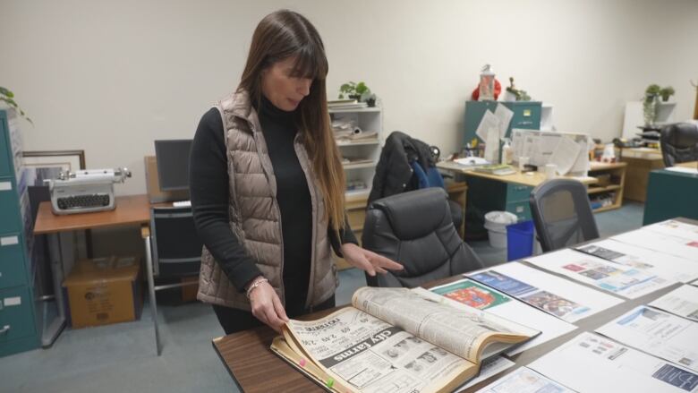 A woman in a vest flips through newspaper archives in an office.