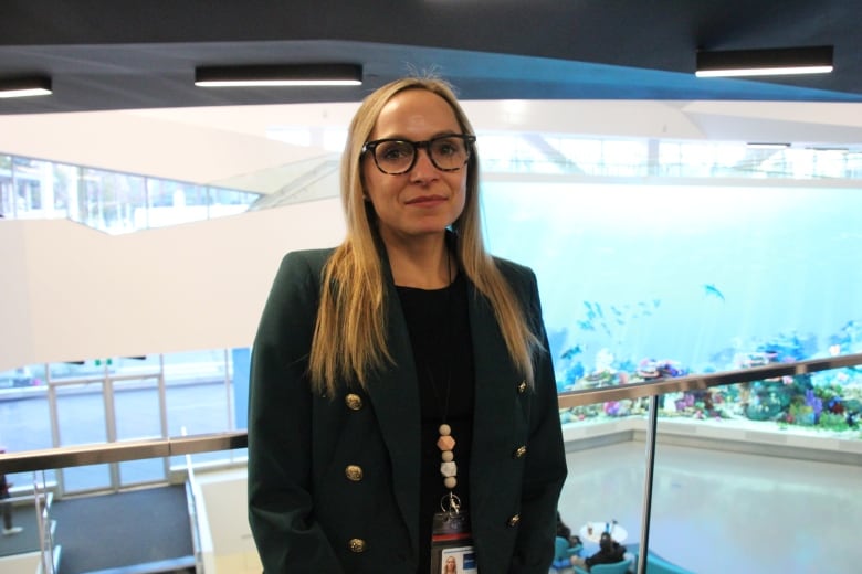 Blonde-haired woman wearing a green blazer stands near a railing overlooking the entrance of the library. A large screen displaying an aquarium takes up the wall behind her.