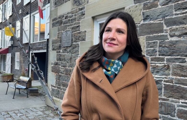 A woman stands in front of a brick building during an interview. She wears a brown coat and a patterned scarf. 