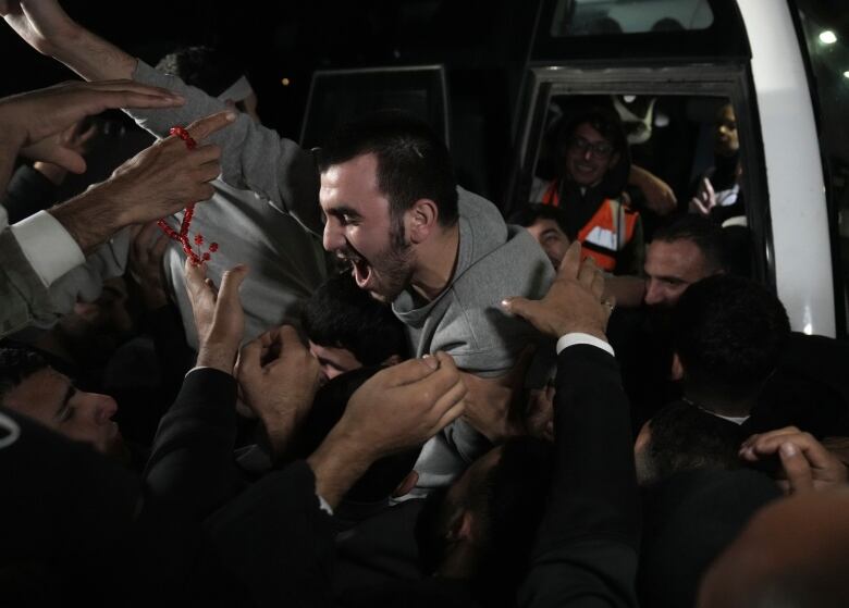 A man, shouting and smiling with one fist pumped in the air, steps out of a vehicle, surrounded by a crowd of people reaching towards him. 