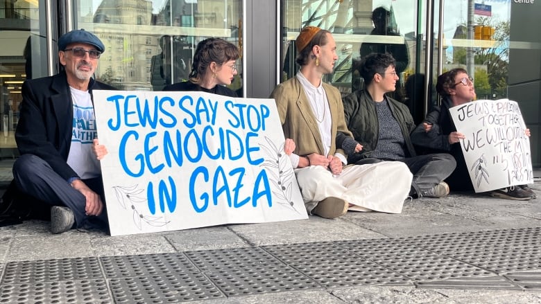 Five people sit cross-legged on a sidewalk, holding placards outside a large building.