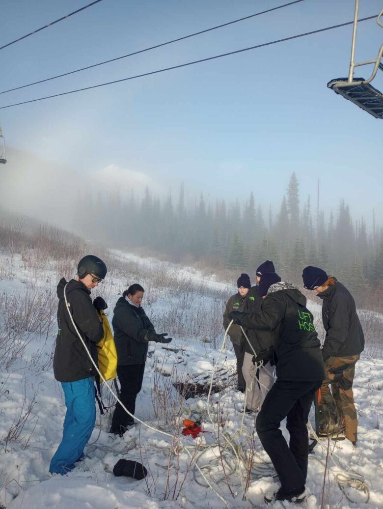 Five people are pictured standing underneath a chairlift on a ski hill, with trees and vegetation visible through the snow.