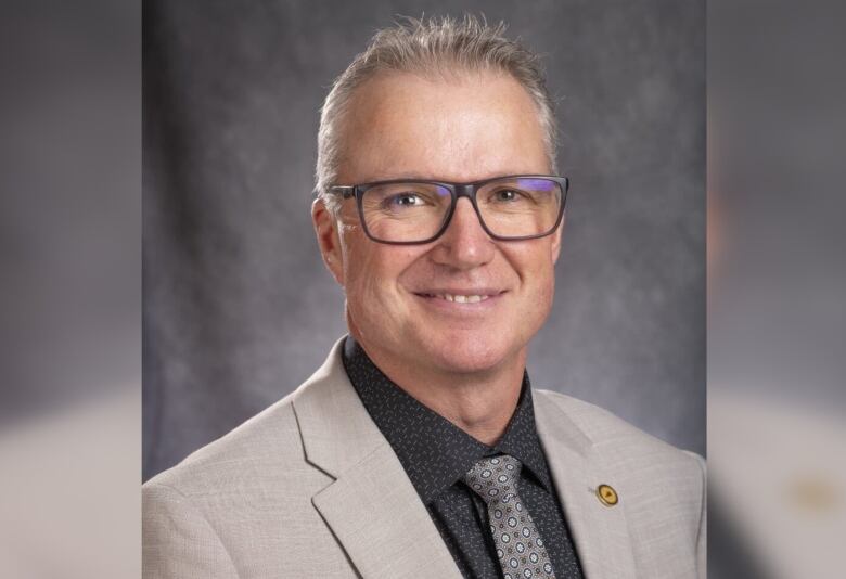 A man has short grey hair and glasses, and wears tan-coloured suit with a dark shirt underneath. He smiles at the camera in a studio portrait.