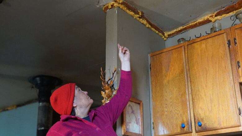 A woman wearing a red knit hat points up toward spray foam that fills a gap between her wall and ceiling above kitchen cabinets. 