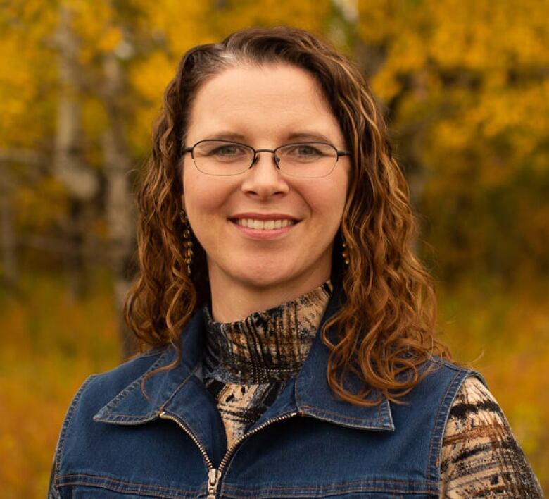 A woman wearing a denim vest over a patterned shirt is pictured with fall foliage in the background.