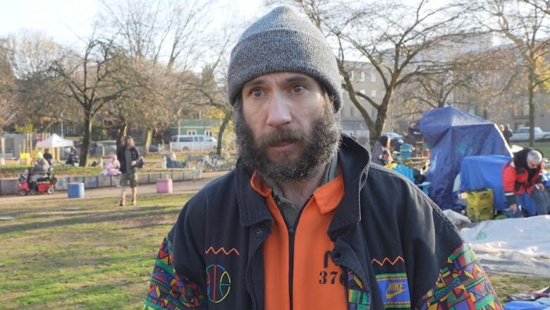 A man with a beard talks against a backdrop of tarps and tents in a city park.