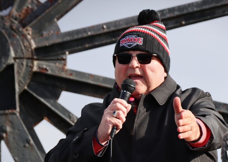 A man with dark glasses, a black parka and a black toque with red and white stripes speaks into a microphone outdoors in front of a large wheel.