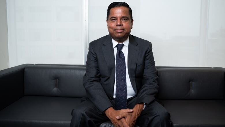 A man in a suit seated on a leather couch with hands folded between his knees.