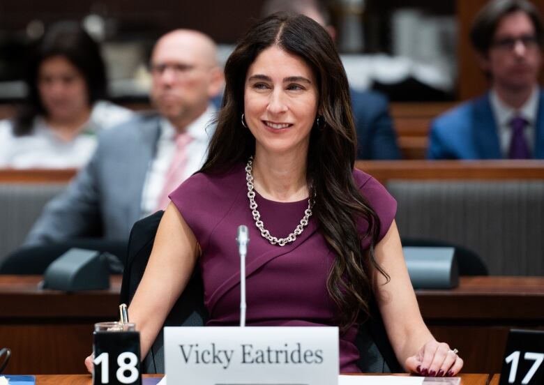 A woman with long brown hair wearing a purple dress and a necklace is seated in parliament.