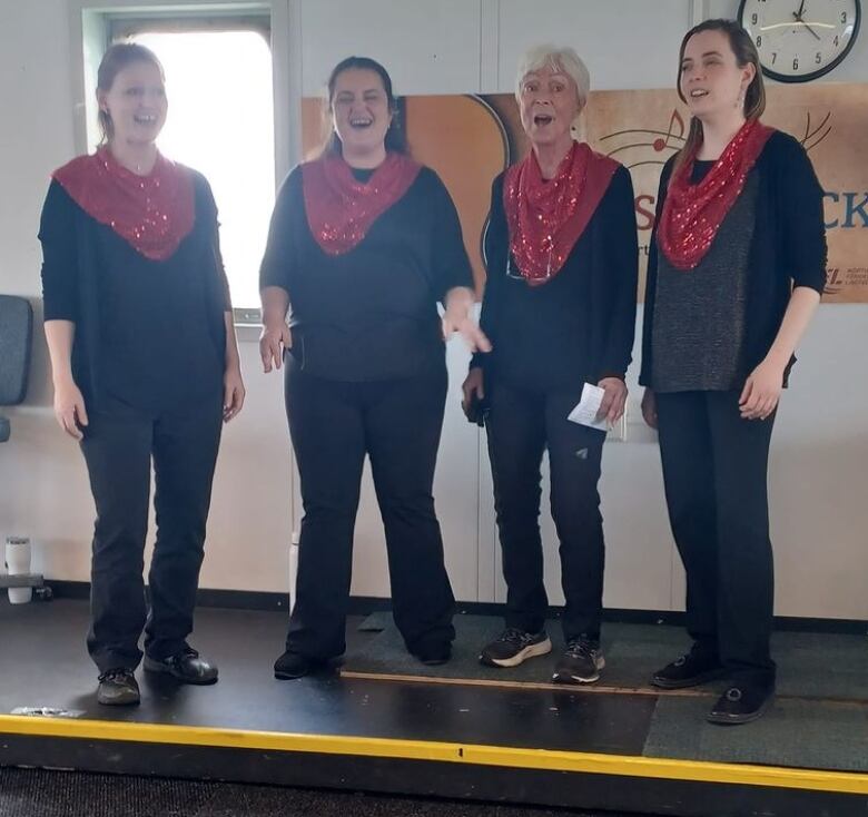 Four women in black clothing and matching red sparkly scarves sing in front of a row of portholes. 