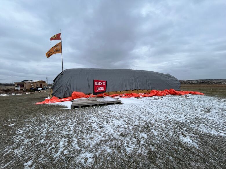 A large wigwam covered with a grey tarp is pictured in an open field on an overcast day with traces of snow on the ground and a sign that reads 'search the landfill.'