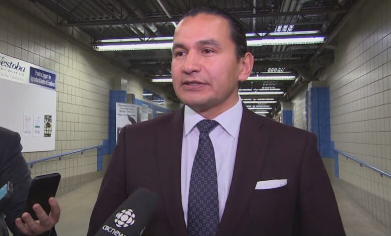 A man wearing a suit jacket, dress shirt and tie stands in front of reporters holding microphones and phone recording devices.