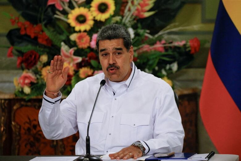 A man with a mustache lifts his arm while speaking in front of a floral backdrop with a flag nearby.