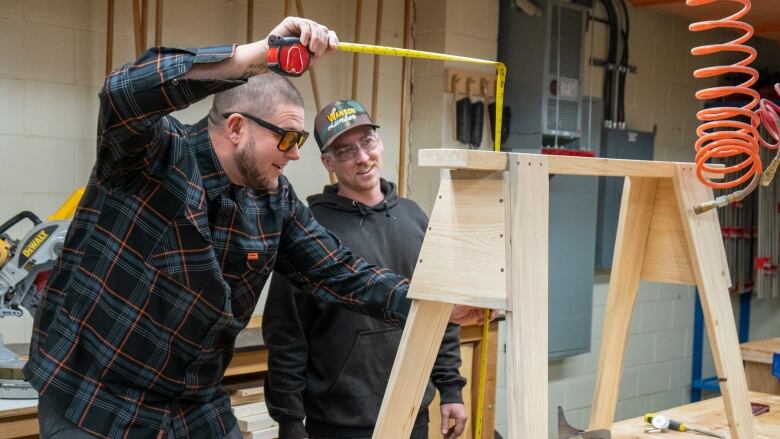 Two people measure a wooden bench in a woodworking shop.