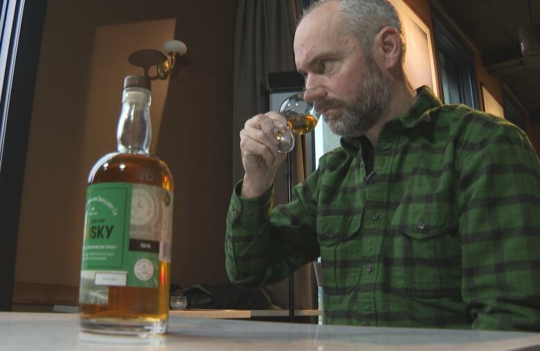 A man in a green, plaid shirt holds a glass of whisky to his nose, sniffing it, a bottle on the table in front of him.