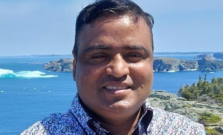 A man smiles at the camera with a rocky shoreline in the background.