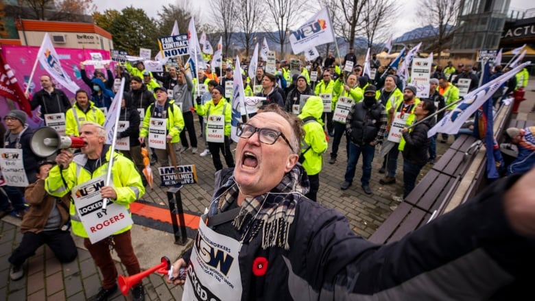 Workers wearing high-visibility vests yell during a protest amid a work stoppage, with many of them wearing sandwich boards saying 'USW Locked out by Rogers'.