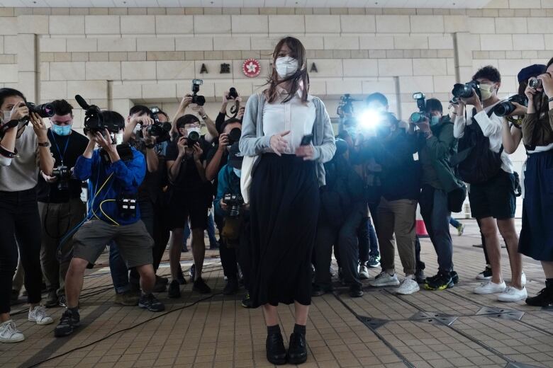 A woman in a face mask is half-encircled by a phalanx of photographers and videographers capturing images of her.