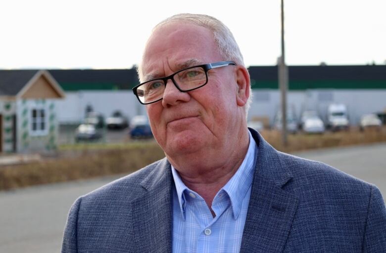 A Caucasian man in glasses and a blazer stands on a street. Behind him is a half-completed building.