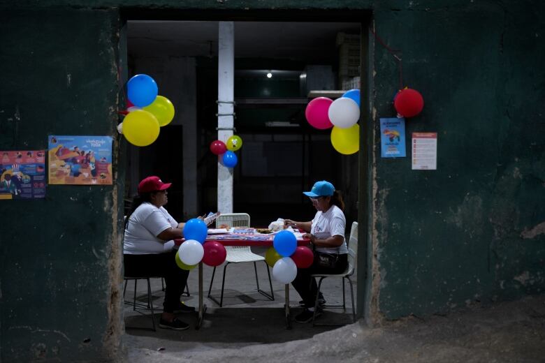 Two people, one in a red hat and one in a blue hat, sit at a table at a polling station.