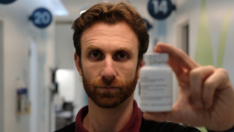 A man with wavy hair and a beard, with a serious expression on his face, holds up a pill bottle.