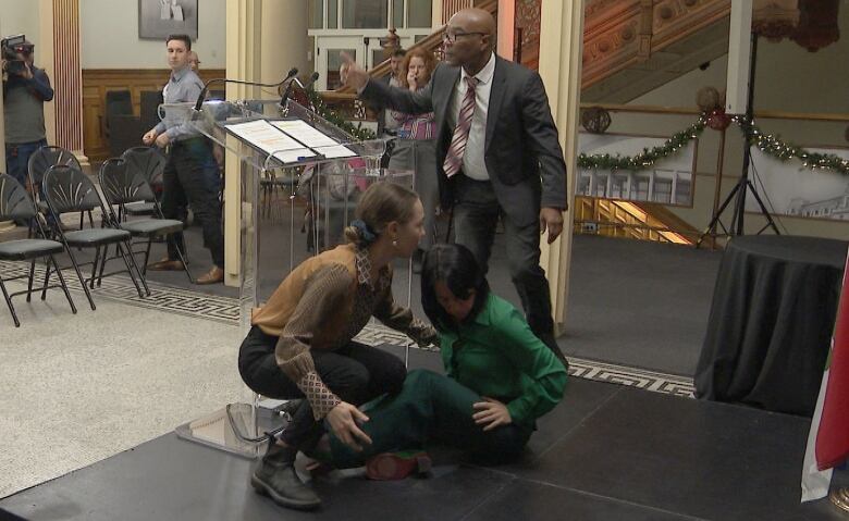 A woman is sitting on a small stage, with another woman crouching over her and a man waving nearby.