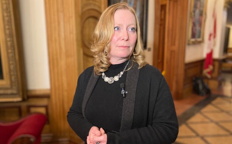 Woman with shoulder-length blonde hair wearing charcoal coloured sweater and cardigan stands inside legislative building.