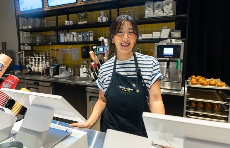 A young woman smiles at the camera from behind a cafe counter. She is visible from the waist up. She's wearing a white t-shirt and apron. 