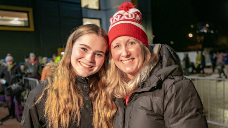 A blonde female teenager with long hair and a black jacket leans against the shoulder of a middle-aged blonde woman also wearing a black jacket as well as a red and white toque that says Canada on it.