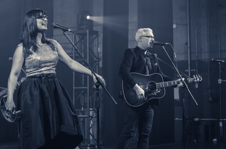 A woman and man sing into microphones on stage. She has long dark hair and is in a sparkly sleeveless top and long black skirt and is holding a microphone. He's in a dark jacket and pants, has short white hair and is playing a guitar.