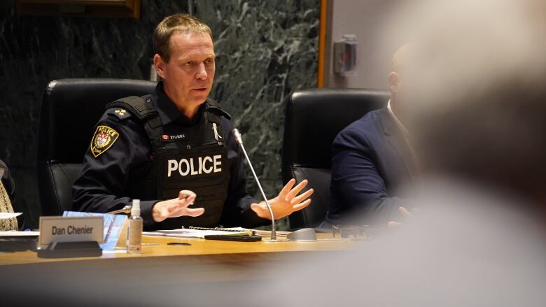 A man in a police uniform sits at a table and speaks into a microphone.