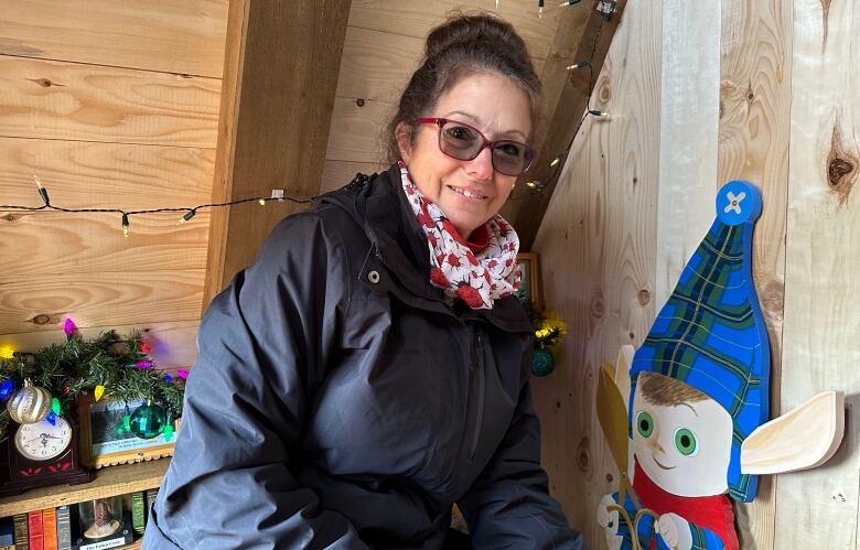 In a chalet decorated for Christmas, a woman wearing winter clothes squats next to a wooden cut out of elf holding tree pruning sheers. 