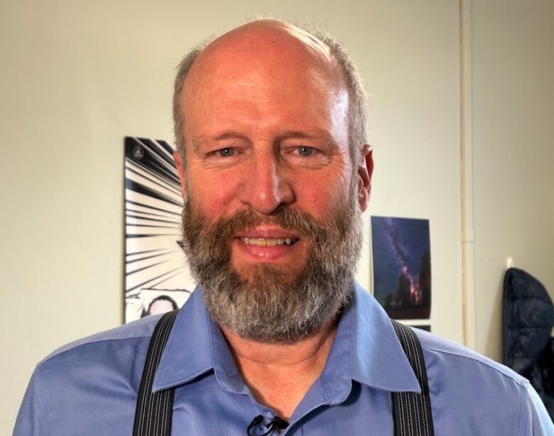 A bald man in a blue shirt and suspenders smiles at the camera.