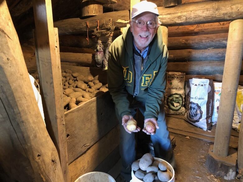 An old white man in a potato cellar holds up two potatoes while smiling.