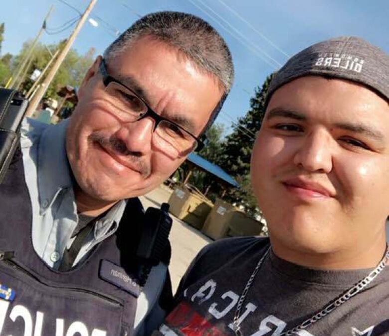 A man wearing a police vest poses for a photo with a younger man wearing a backwards ball cap. 