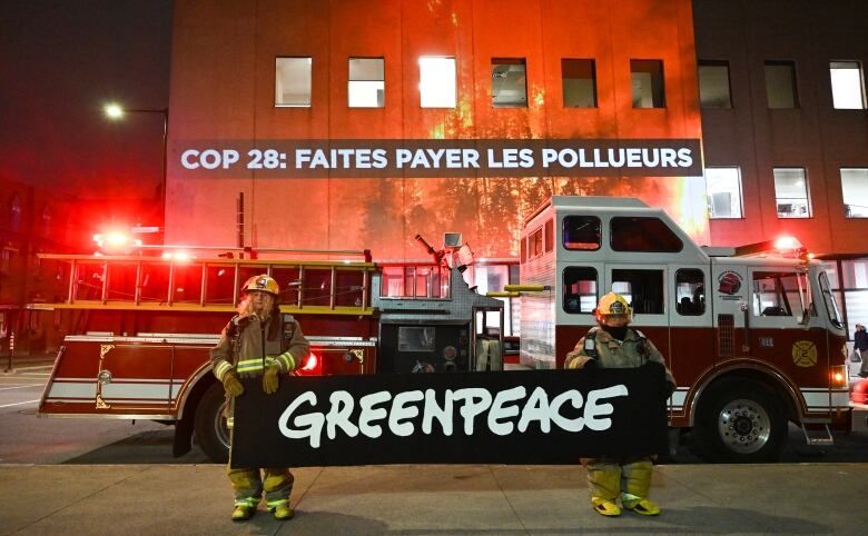 Two people dressed as firefighters hold up a Greenpeace banner while standing in front of a fire truck and a building with the image if a forest fire projected onto it. The projection also includes the words: 'COP28: FAITES PAYER LES POLLUERS.'