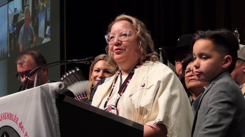 A First Nations woman surrounded by people, wearing white buckskins speaks into a microphone.