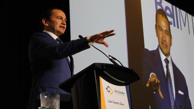 Man standing at podium holds out his right hand, while an image of him is projected in the background.