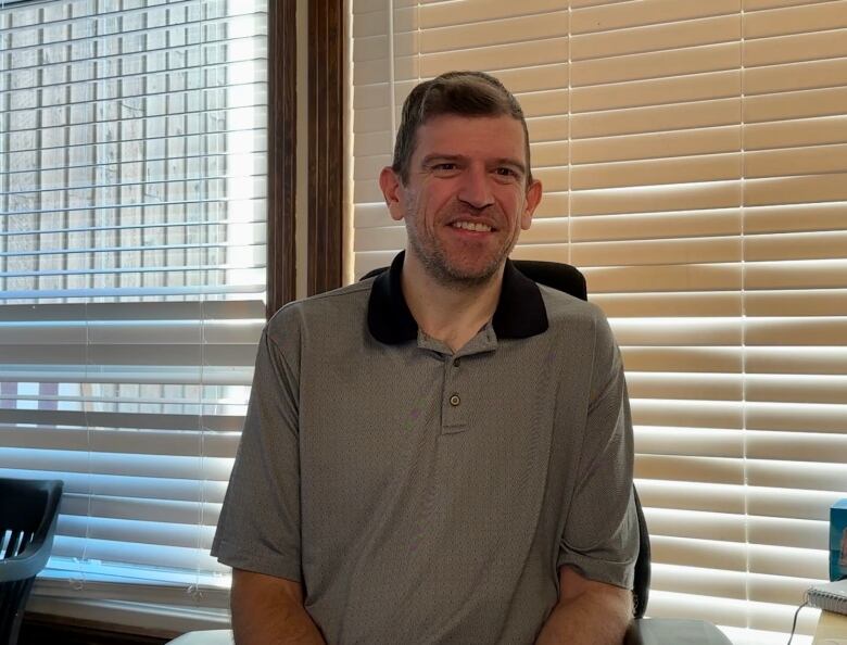 A smiling caucasian man with brown hair seated in front of a window, wearing a gray golf shirt with a black collar. 