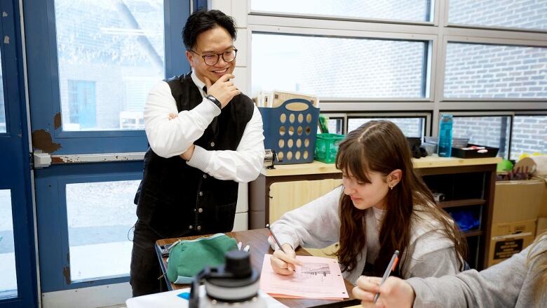 A man smiles as he stands beside students working at tables in a classroom.